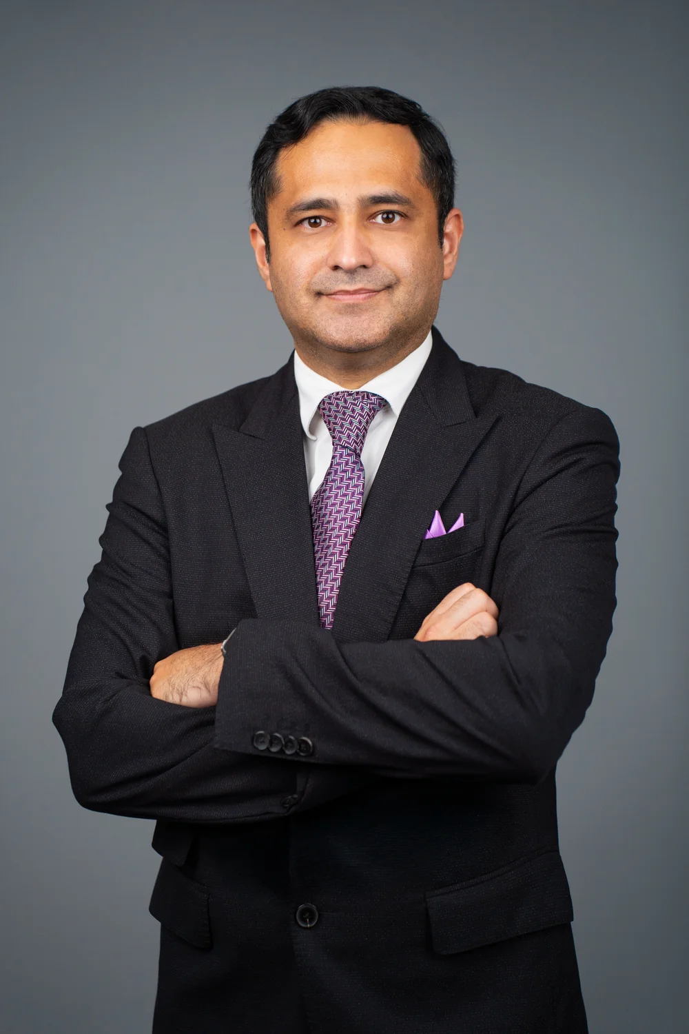 Zahaib Nabeel in a dark suit and tie with arms crossed, photographed against a grey studio background